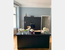 Moderne Küche mit einer dunkelgrauen Kücheninsel aus Stein und darauf mehrere Blumenvasen mit weißen und farbigen Blumen. Im Hintergrund ein hoher, dunkelgrauer Einbauschrank mit einem eingebauten Backofen, darüber eine Buddha-Statue. Der Raum hat helle Wände, Parkettboden und Tageslicht von einem großen Fenster links.