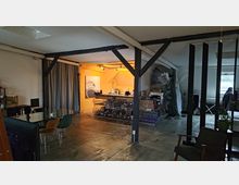 Großzügiger Raum in einer industriellen Loft-Ästhetik mit Betonboden, sichtbaren Stahlträgern und gedeckter Beleuchtung. Links befindet sich ein Bereich mit Glastisch und Stühlen, rechts eine offene Ecke mit Regalen und gelagerten Gegenständen; im Hintergrund ein kleiner Küchenbereich mit gelbem Licht.