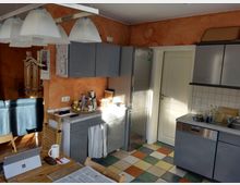 Küche mit grauen Schränken, einer Edelstahl-Kühlschranktür und einer weißen Tür im Hintergrund. Der Boden besteht aus bunt gemusterten Fliesen, und auf der Arbeitsplatte stehen Haushaltsgegenstände wie Gläser und Gewürze; im Vordergrund befindet sich ein Holztisch mit Büromaterial.