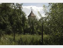 Ein Turm mit spitzem Dach und Wetterfahne ragt hinter dichtem Grün eines Gartens hervor, der von einem Drahtzaun umgeben ist. Der Himmel ist leicht bewölkt, und die Vegetation besteht aus hohen Bäumen und Sträuchern.