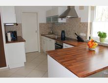 Moderne Einbauküche mit weißen Schränken, Arbeitsplatten aus Holzoptik und beigem Fliesenspiegel. Ausgestattet mit einer Dunstabzugshaube aus Edelstahl, einem Spülbecken, einem Herd sowie einem Obstkorb und Basilikumtopf auf der Theke.