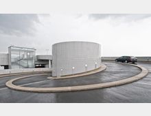 Eine spiralförmige Auffahrtsrampe aus Beton in einem Parkhaus, mit einem zylindrischen Treppenturm aus Glas und Metall auf der linken Seite. Ein dunkles Auto fährt auf der asphaltierten Rampe, und der Himmel ist bewölkt.