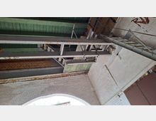 Blick nach oben in einen industriellen Innenraum mit weiß gestrichenen Ziegelwänden und einem Rundbogen, darüber grün gestrichene Holzdecke. Rostige Stahlträger, Metallstreben und eine Leiterkonstruktion verlaufen entlang der Wand, daneben ist ein großes Metalltor zu sehen.