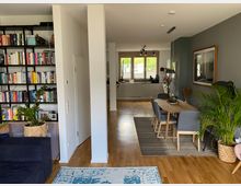 Ein modernes Wohnzimmer mit offener Küche im Hintergrund. Links steht ein schwarzes Bücherregal, rechts ein Essbereich mit Holztisch, grauen Stühlen und Dekorationen, umgeben von Zimmerpflanzen.