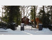 Holzhaus in winterlicher Umgebung, umgeben von verschneiten Bäumen und Sträuchern. Im Vordergrund ein schmiedeeisernes Tor mit zwei Steinsäulen, daneben eine überdachte Holzstruktur.