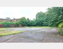 Ein leerer, betonierter Platz mit vereinzeltem Pflanzenbewuchs, umgeben von dichten Bäumen und Sträuchern. Im Hintergrund steht ein längliches, backsteinernes Gebäude mit mehreren Fenstern und einem Zaun davor.