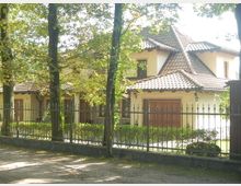 Einfamilienhaus im mediterranen Stil mit cremefarbener Fassade, roten Dachziegeln und Holzfenstern, umgeben von einem Metallzaun und Bäumen im Vordergrund. Der gepflegte Garten ist teilweise sichtbar.
