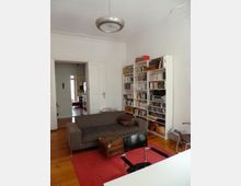 Wohnzimmer mit Holzboden, grauem Sofa und rotem Teppich. Im Hintergrund stehen ein Bücherregal und ein Esstisch, links führt eine weiße Doppeltür in einen angrenzenden Raum mit Küche.