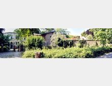 Eine idyllische Außenansicht mit einem Herrenhaus in neoklassizistischem Stil links im Bild, einer Backsteingebäude rechts daneben und einer alten Steinmauer im Vordergrund, umgeben von dichtem Grün, Sträuchern und Bäumen. Ein gepflasterter Weg führt zu einem schmiedeeisernen Tor.