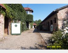 Hof mit Kopfsteinpflasterweg, flankiert von Backsteingebäuden; links ein mit Efeu bewachsenes Gebäude mit grünen Stalltoren, rechts ein langes Gebäude mit offenen Rundbögen. Im Hintergrund sind weitere Hofstrukturen und Bäume sichtbar.