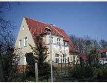 Zweistöckiges Wohnhaus mit gelber Fassade, Satteldach aus roten Ziegeln und mehreren weißen Fenstern, umgeben von einem Garten mit Zaun. Im Hintergrund sind Bäume und ein klarer blauer Himmel zu sehen.