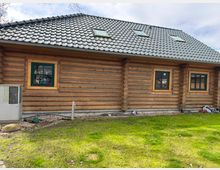 Außenansicht eines Blockhauses mit Wänden aus natürlichen Holzstämmen, grünen Fenstern mit Rahmen und einem grauen Ziegeldach mit Dachfenstern. Im Vordergrund befindet sich ein gepflegter Rasen und eine kleine technische Anlage links am Haus.