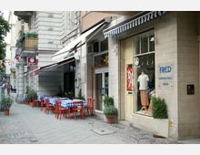 Straßenszene mit Geschäftsfassaden, darunter eine Kinderboutique mit dem Namen „FRED“ und ein Restaurant mit rot gedeckten Tischen und blau-weiß karierten Tischdecken auf dem Gehweg. Die Umgebung besteht aus historischen Gebäuden mit Balkonen und dekorativen Details, flankiert von kleinen Pflanzen in Töpfen.