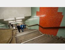 Treppenabgang in einer U-Bahn-Station mit beige-grauen Stufen und Handläufen an beiden Seiten. Die Wände sind mit roten und blauen Fliesen gestaltet, im Hintergrund befindet sich ein Schild mit den Linien U9 und U3.