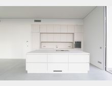 Moderne, minimalistische Küche in weißem Design mit einer großen Kücheninsel, glatten Schränken und integrierten Geräten, darunter ein Backofen. Die Wände und Böden sind schlicht gestaltet, helle Farben dominieren den Raum.