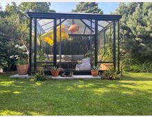 Schwarzes, verglastes Gartenhaus mit Satteldach in einem grünen Garten, umgeben von Bäumen und Sträuchern. Innen sind eine L-förmige Sitzgruppe und eine Hängelampe zu sehen, davor stehen mehrere Terrakotta-Blumentöpfe auf einer niedrigen Steinplatte.