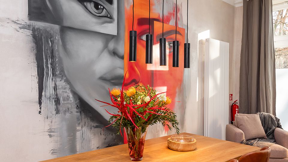 Moderner Essbereich mit großem Holztisch und braunen Lederstühlen unter einer Hängelampe mit mehreren schwarzen Pendelleuchten. An der Wand befindet sich ein großflächiges Graffiti-Porträt, daneben ein Fenster mit Vorhängen sowie ein Heizkörper und ein Sofa in der Ecke.