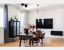 Essbereich in einem modernen Wohnzimmer mit hellem Holzfußboden, einem rechteckigen Holztisch und gemischten Stühlen. An der Wand hängt ein großer Flachbildfernseher, daneben ein schwarzer Schrank mit Glasfront; Fenster mit weißen und grauen Vorhängen lassen Tageslicht herein.