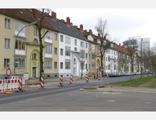 Straßenszene mit mehrstöckigen Wohnhäusern aus der Nachkriegszeit, die verputzt und in hellen Beige- und Weißtönen gehalten sind. Im Vordergrund befinden sich eine Fahrbahn mit provisorischen Absperrungen und Hinweisschildern sowie einige geparkte Autos am Straßenrand; kahle Bäume säumen die Straße.