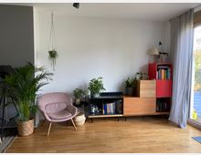 Wohnzimmer mit hellen Holzböden, einer gepolsterten rosa Sitzgelegenheit, mehreren Zimmerpflanzen in Körben und Töpfen, einer minimalistischen Regal-Stapelung in Rot- und Holztönen sowie einem großen Fenster mit grauen Vorhängen.