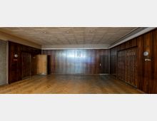 Holzvertäfelter Raum mit Parkettboden und abgehängter, rechteckig gemusterter Decke. Die Wände bestehen aus dunklem Holz mit Türen, einer offenen und mehreren geschlossenen; ein Wappen hängt zentral an der rückseitigen Wand.