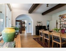 Esszimmer in einem hellen Wohnhaus mit Holzboden, Esstisch und Holzstühlen auf einem bunten Teppich. Im Raum befinden sich außerdem ein Klavier, ein Bücherregal und große Fenster; ein bogenförmiger Durchgang führt zu weiteren Räumen.