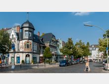 Straßenszene mit historischen Gebäuden, darunter ein verziertes Fachwerkhaus mit einem Turm und bemalter Fassade. Im Vordergrund eine gepflasterte Straße mit geparkten Autos und Fußgängern, umgeben von Bäumen und Ladengeschäften.