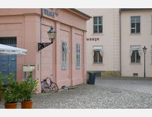 Rosa Gebäude mit weißgerahmten Fenstern und einer blauen Tür auf einem gepflasterten Platz; daneben steht ein Fahrrad, sowie eine Laterne an der Fassade. Im Hintergrund helles Gebäude mit Fenstern und heruntergelassenen Vorhängen.