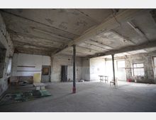 Großer, leerer Industrie- oder Lagerraum mit bröckelnder Decke, freiliegenden Trägern und Stützsäulen sowie alten Neonröhren. An den abgenutzten Wänden stehen vereinzelte Möbel und eine Leiter; Tageslicht fällt durch mehrere Fenster auf den Betonboden.