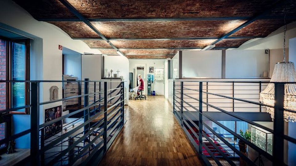 Innenansicht eines modernen Loftbüros mit langem Holzfußboden, Metallgeländern entlang eines offenen Galeriegangs und sichtbarer Ziegeldecke. Links sind große Fenster und Arbeitsbereiche zu sehen, rechts öffnet sich der Raum zu einer tieferliegenden Ebene mit Treppe und hängender Leuchte.