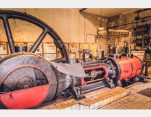 Industriehalle mit einer historischen Dampfmaschine, bestehend aus einem großen schwarzen Schwungrad und einem roten Zylinder. Im Hintergrund sind alte Schaltkästen, Rohre und bröckelnde Wände sichtbar.