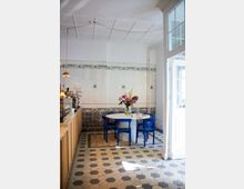 Heller Innenraum eines Cafés mit hexagonalem Fliesenboden, einer weißen gefliesten Wand mit Ziermuster und einem runden Tisch mit blauer Bestuhlung. Auf dem Tisch steht ein Blumenstrauß in einer Vase, rechts neben der Szene befindet sich eine große weiße Glastür.