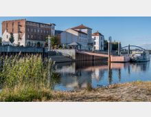 Stadtansicht mit Uferpromenade, historischen Gebäuden aus Backstein und Putzfassaden, einer kleinen Fußgängerbrücke und einem bewachsenen Uferbereich im Vordergrund; rechts ein angelegtes Boot am Wasser.