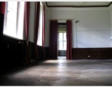 Ein leerer Raum in einem historischen Gebäude mit hohen Decken, dunklen Holzwandverkleidungen und großen Fenstern, die von roten Vorhängen eingerahmt sind. Der Boden besteht aus Holzparkett, und an einer Wand sind Risse sichtbar.