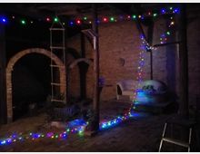 Ein rustikaler Innenhof mit Backsteinwänden und Rundbögen, dekoriert mit bunten Lichterketten. Im Vordergrund steht eine Holzleiter, daneben befinden sich ein Sofa mit Decken und Pflanzen in Töpfen.