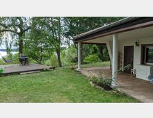 Eine Terrasse mit Fliesenboden und weißen Säulen, angrenzend an einen Garten mit Rasen und Bäumen. Im Hintergrund ist ein hölzernes Podest mit einem Tisch, Stühlen und einer Hängematte zu sehen, sowie ein Blick auf einen See.