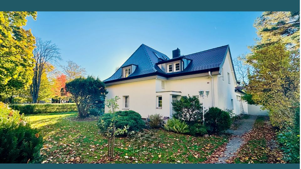 Außenansicht eines hellen Einfamilienhauses mit dunklem Satteldach und Gauben, umgeben von einem großen Garten mit Rasen, Sträuchern und hohen Bäumen. Ein schmaler Weg führt am Haus entlang, herbstliches Laub liegt auf dem Boden.