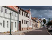 Straße mit Reihenhäusern in historischem Baustil, teils mit Fachwerk und verputzten Fassaden. Der Gehweg ist gepflastert, Laternen und parkende Autos säumen die Straße, während dunkle Wolken den Himmel bedecken.
