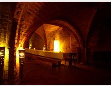 Gewölbekeller aus Ziegelsteinen mit beleuchteten Rundbögen, einem langen, weiß gedeckten Tisch und stimmungsvoller Lichtgestaltung durch Laternen und warme Lampen. Der Boden besteht aus rot-braunen Fliesen und einfache Stühle ergänzen das Ambiente.