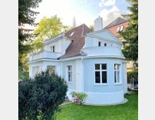 Zweistöckiges Haus mit hellem Putz und rundem Erker, roten Ziegeldach und großen Fenstern, umgeben von Garten mit Rasen, Büschen und Bäumen bei sonnigem Wetter.