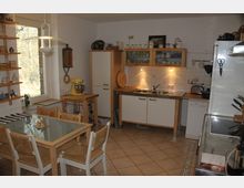 Helle Küche mit Holz- und Weiß-Akzenten, bestehend aus einem Esstisch mit vier Stühlen, einem Herd, einem Kühlschrank und einer Spüle. An den Wänden befinden sich Regale mit Geschirr und Dekorationen, ein großes Fenster bietet Blick auf eine baumreiche Außenumgebung.