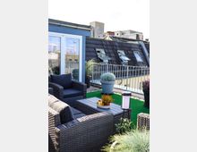 Eine moderne Dachterrasse mit Sitzmöbeln aus grauem Flechtmaterial, einem niedrigen Tisch und großer Pflanzendekoration. Im Hintergrund ist ein Hausdach mit Dachfenstern sowie der Ausblick auf städtische Gebäude sichtbar.