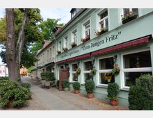 Straßenszene mit einer grünen, zweistöckigen Fassade eines Gasthauses mit der Aufschrift „Gast- und Logierhaus Zum Jungen Fritz“. Im Vordergrund befinden sich Gehwegplatten, Blumenkästen an den Fenstern sowie Sitzgelegenheiten vor dem Gebäude, umgeben von Bäumen und Sträuchern.