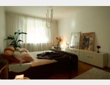 Gemütlich eingerichtetes Schlafzimmer mit einem Polsterbett, weichen Kissen und einer Decke. Im Hintergrund ein Fenster mit transparenten Vorhängen, eine Kommode mit dekorativen Vasen, Kerzen und gerahmten Bildern, sowie eine Zimmerpflanze in der Ecke.