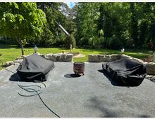 Gartenbereich mit einer gepflasterten Terrasse, auf der zwei mit schwarzen Abdeckungen geschützte Möbel stehen. Im Hintergrund eine Wiese mit einem Brunnen, Statuen und dichtem Baumbewuchs; eine Steinmauer und Laternen grenzen die Terrasse ab.
