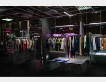 Ein moderner Bekleidungsladen mit Metallständern, auf denen bunte Kleidungsstücke hängen, und einer zentralen Tischfläche. Der Raum hat eine dunkle Atmosphäre mit Neonlichtern und großen Fensterfronten, die den Blick nach draußen ermöglichen.