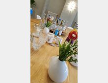 Ein großer Holztisch mit verschiedenen Trinkflaschen, Gläsern, Tassen und kleinen weißen Vasen mit Blumen. Im Hintergrund sind eine Wandheizung, weiße Stühle und eine Wandleuchte sichtbar.