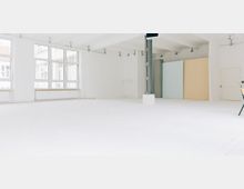 Heller, leerer Innenraum mit weißen Wänden und Boden, großen rechteckigen Fenstern an der linken Seite und einer Trägerstruktur in der Mitte. Im Hintergrund befinden sich pastellfarbene Trennwände in Beige und Hellblau sowie eine weiße Tür.