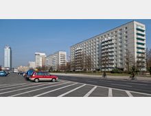 Straßenansicht mit Mehrfamilienhäusern im Plattenbaustil, flankiert von Bäumen und einer parkähnlichen Grünfläche. Im Vordergrund ist eine breite Straße mit parkenden Autos und Radfahrern zu sehen; im Hintergrund steht ein Hochhaus.