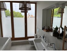 Modernes Badezimmer mit großen, bodentiefen Fenstern, die durch Milchglas teilweise blickdicht sind. Zu sehen sind ein Doppelwaschbecken mit Spiegel, eine Badewanne, ein WC sowie dekorative Elemente wie Pflanzen und Hängeleuchten aus geflochtenem Material.